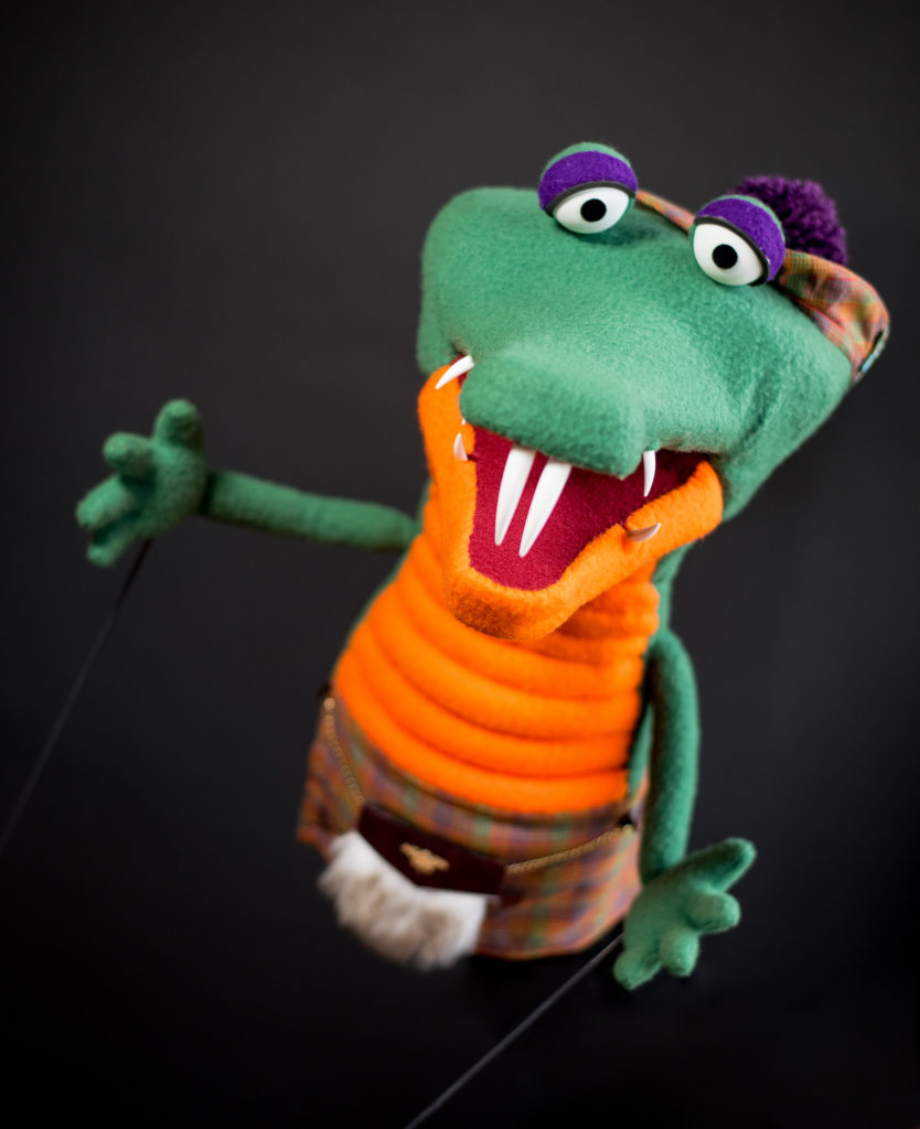 Angus, a Green and orange Scottish crocodile, wearing a plaid kilt, tam, and fur sporran.