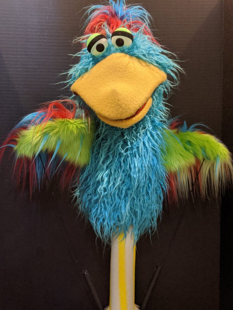 Blue bird puppet with big yellow beak, big eyes, and muticolored wings and top feathers.