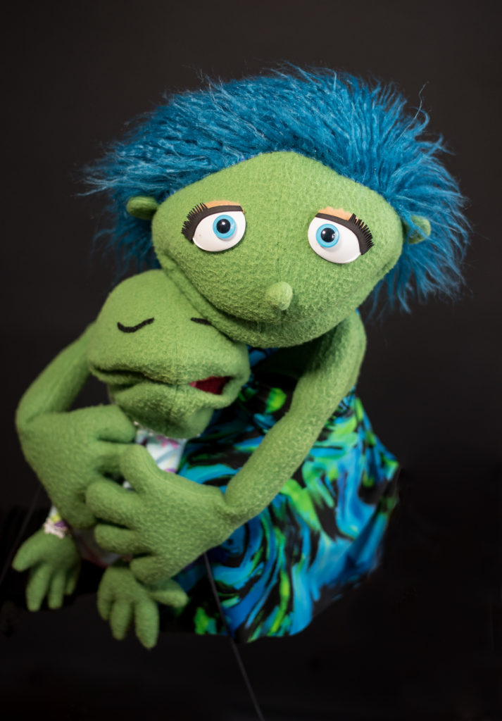 Mother puppet with green skin and blue hair is holding a baby puppet with green skin.