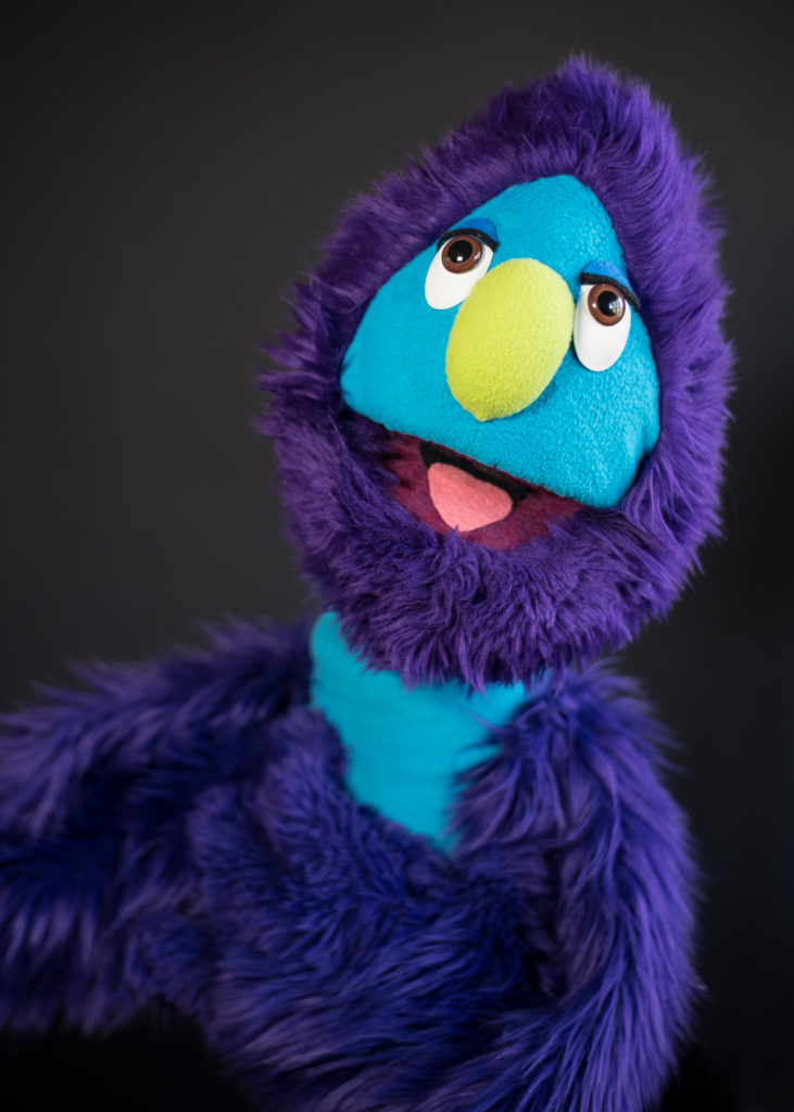 Live hand puppet with purple furry head and arms, blue skin, and a big green nose.