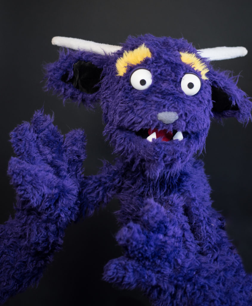 Live hand purple furry monster puppet with horns, lower fangs, big white eyes, and yellow eyebrows.